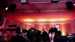 fourt people sit in a row on stage. one is holding a microphone and speaking as part of a panel.