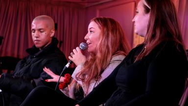 a trio of people sit on stage, one is holding a microphone and speaking