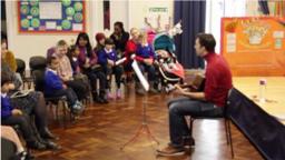 a person sitting down with a guitar, playing in front of a crowd of children and parents in a school classroom