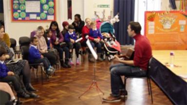 a person sitting down with a guitar, playing in front of a crowd of children and parents in a school classroom