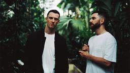 two men in white t-shirts, one wearing a black jacket, standing in a jungle 