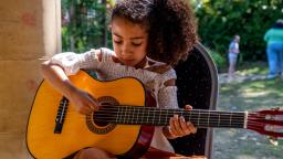 A young musician sat down playing a guitar with some trees behind them