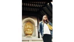 Person with long brown hair, wearing a denim jacket, white shirt, and black trousers standing in front of a golden statue.