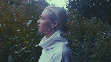girl wearing white jumper with hair in a bun from a side profile, trees and green plants behind her