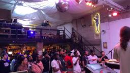 a crowd of people stand in a purple lit room and face the stage. a disco ball hangs overhead