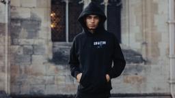 young person wearing a black hoody with hands in their pockets, brick building with windows behind them