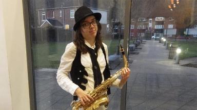 person wearing white shirt and black waistcoat holding a saxaphone. Standing in front of a glass door, houses on a street visible
