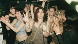 a group of young women cheer and clap in a room. one woman in the centre is holding a microphone