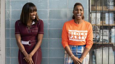 Two black people standing and laughing. One is wearing a burgundy dress, the other is wearing a orange jumper and patterned trousers.