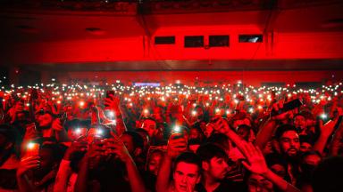a crowd in a music venue all pointing their phones towards the camera, bathed in red light