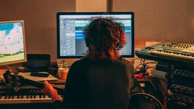 back of person sitting at computer with electric keyboards visible 