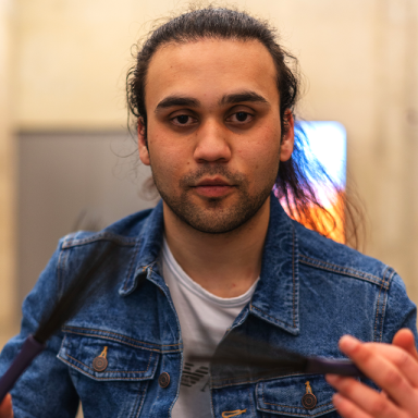 a man with dark hair wears a white t-shirt and blue denim jacket