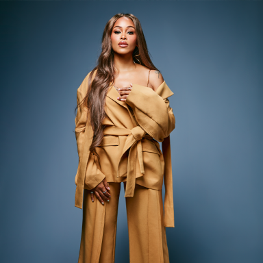 a woman with long brown hair poses with one hand on her chest. she is wearing a draped brown material