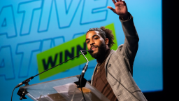 superlative on stage after winning an award, his hand is raised in the air and he wears a grey tweet suit