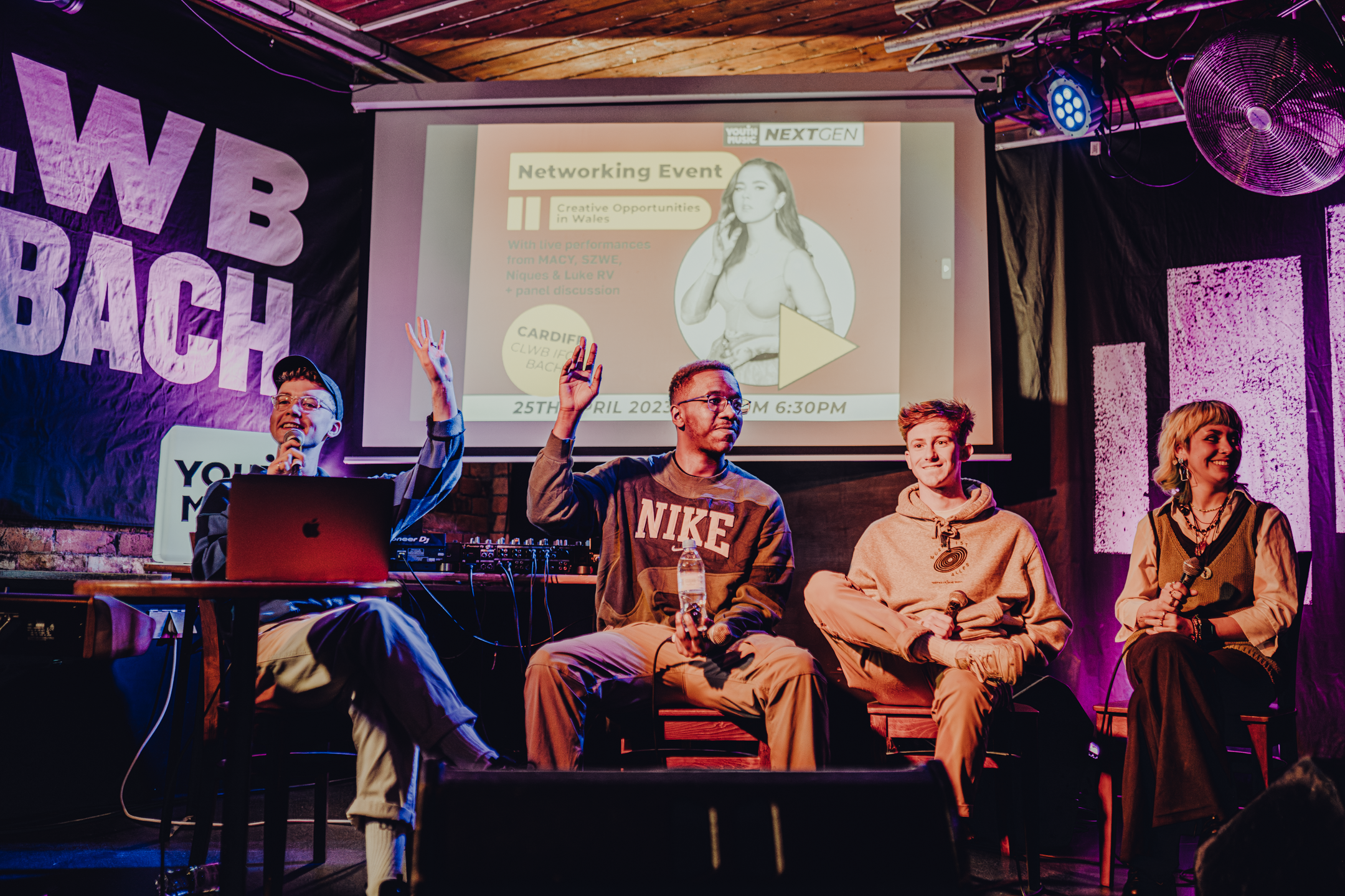 four young people sit on chairs on a stage as part of a panel. they are smiling, raising their hands and hold microphones